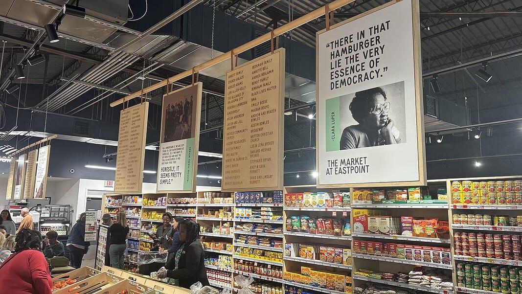 Interior of a grocery or market with hanging posters above long aisles and shoppers browsing shelves of packaged foods.