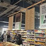 Interior of a grocery or market with hanging posters above long aisles and shoppers browsing shelves of packaged foods.
