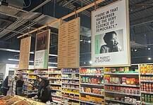 Interior of a grocery or market with hanging posters above long aisles and shoppers browsing shelves of packaged foods.