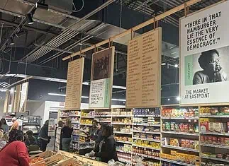 Market at EastPoint marks 5 years amid tough economy, Homeland plans Interior of a grocery or market with hanging posters above long aisles and shoppers browsing shelves of packaged foods.