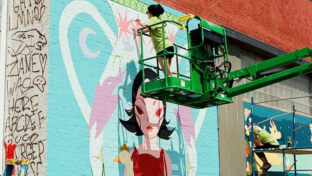 Two workers on a green boom lift painting a colorful mural of a woman with black hair on a brick wall.