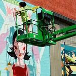 Two workers on a green boom lift painting a colorful mural of a woman with black hair on a brick wall.