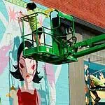 Sunny Dayz Mural Fest heads to Claremore with inclusive art, OKC artists Two workers on a green boom lift painting a colorful mural of a woman with black hair on a brick wall.