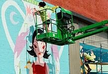 Two workers on a green boom lift painting a colorful mural of a woman with black hair on a brick wall.