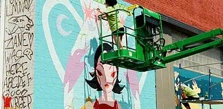 Two workers on a green boom lift painting a colorful mural of a woman with black hair on a brick wall.