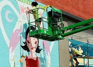 Sunny Dayz Mural Fest heads to Claremore with inclusive art, OKC artists Two workers on a green boom lift painting a colorful mural of a woman with black hair on a brick wall.