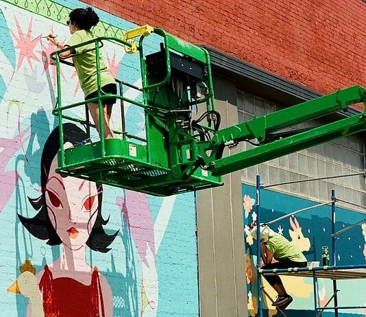 Sunny Dayz Mural Fest heads to Claremore with inclusive art, OKC artists Two workers on a green boom lift painting a colorful mural of a woman with black hair on a brick wall.