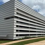 Modern white building with horizontal slat facade and a glass entrance, surrounded by a grassy lawn under a blue sky.
