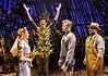Actor in a leafy, harvest-themed costume raises arms on stage as four costumed performers look on with smiles on a wooden theater set.