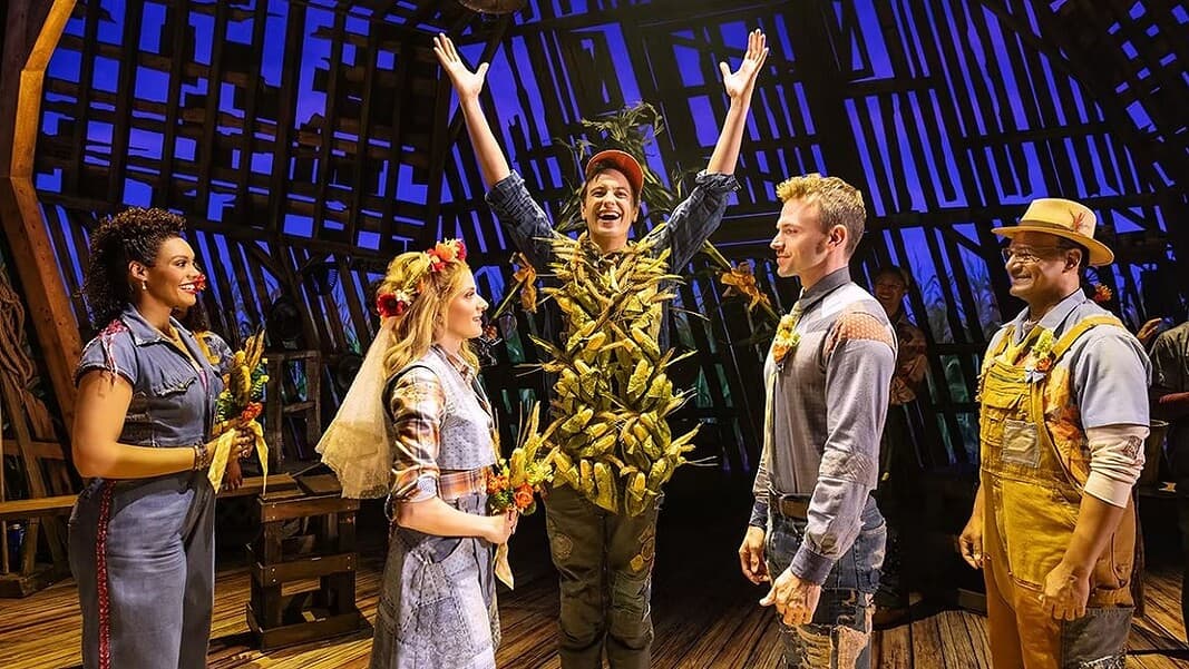 Shucked 5 Actor in a leafy, harvest-themed costume raises arms on stage as four costumed performers look on with smiles on a wooden theater set.