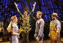 Actor in a leafy, harvest-themed costume raises arms on stage as four costumed performers look on with smiles on a wooden theater set.