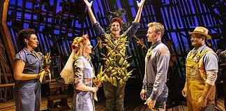 Actor in a leafy, harvest-themed costume raises arms on stage as four costumed performers look on with smiles on a wooden theater set.