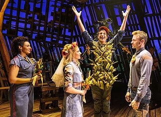 Actor in a leafy, harvest-themed costume raises arms on stage as four costumed performers look on with smiles on a wooden theater set.