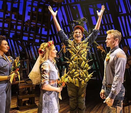 Actor in a leafy, harvest-themed costume raises arms on stage as four costumed performers look on with smiles on a wooden theater set.