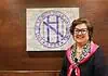 PRESS RELEASE: Nichols Hills elects Sody Clements to serve sixth term as mayor Smiling woman in a black top with a pink scarf stands beside a wall featuring a large circular purple Nichols Hills logo.