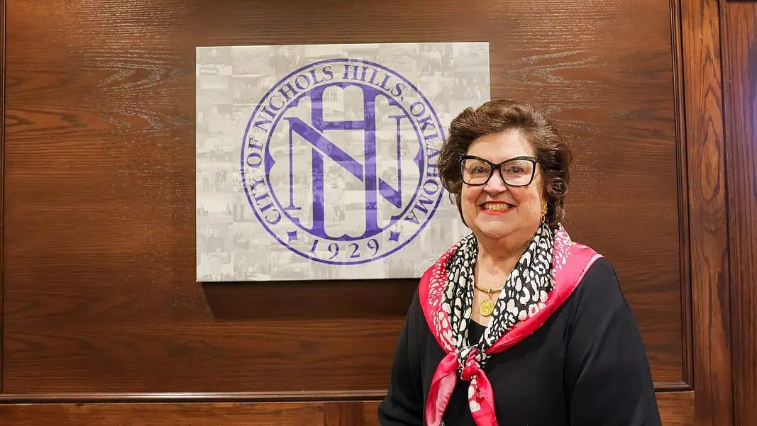 SodyClements2 Smiling woman in a black top with a pink scarf stands beside a wall featuring a large circular purple Nichols Hills logo.