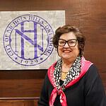 Smiling woman in a black top with a pink scarf stands beside a wall featuring a large circular purple Nichols Hills logo.