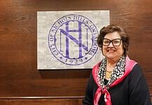 Smiling woman in a black top with a pink scarf stands beside a wall featuring a large circular purple Nichols Hills logo.