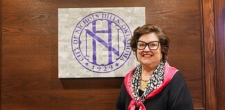 Smiling woman in a black top with a pink scarf stands beside a wall featuring a large circular purple Nichols Hills logo.