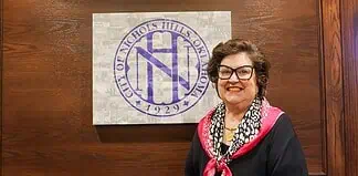 Smiling woman in a black top with a pink scarf stands beside a wall featuring a large circular purple Nichols Hills logo.