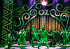 Stage with neon-green 1920s signage and palm-leaf motifs; a group of dancers in green costumes perform a choreographed routine.