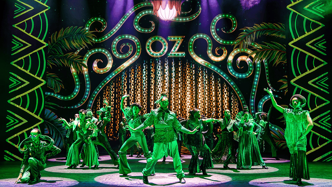 Stage with neon-green 1920s signage and palm-leaf motifs; a group of dancers in green costumes perform a choreographed routine.