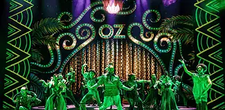 Stage with neon-green 1920s signage and palm-leaf motifs; a group of dancers in green costumes perform a choreographed routine.