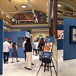 Museum gallery with blue walls, visitors viewing colorful artworks and a pink sculpture in a glass display case on a pedestal.