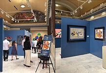 Museum gallery with blue walls, visitors viewing colorful artworks and a pink sculpture in a glass display case on a pedestal.
