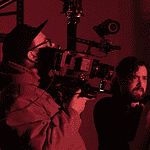 Cameraman operating a large professional camera while a man and woman converse in a red-lit studio space.