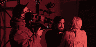 Cameraman operating a large professional camera while a man and woman converse in a red-lit studio space.