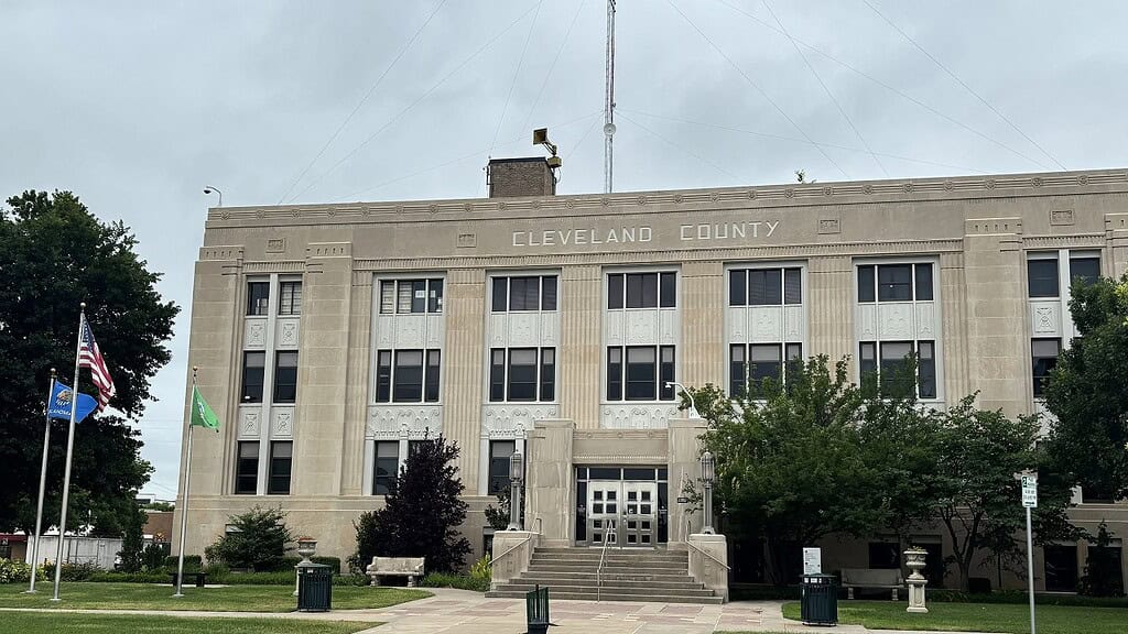 Cleveland County courthouse, a beige stone building with tall windows, flags on poles, and steps leading to the entrance. Trees and benches in front.