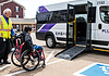 EMBARK driving accessibility, assistance for public transit in OKC Caregiver in a safety vest helps a person in a red wheelchair toward a white shuttle van with a lowered ramp in a parking lot.