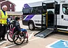 EMBARK driving accessibility, assistance for public transit in OKC Caregiver in a safety vest helps a person in a red wheelchair toward a white shuttle van with a lowered ramp in a parking lot.