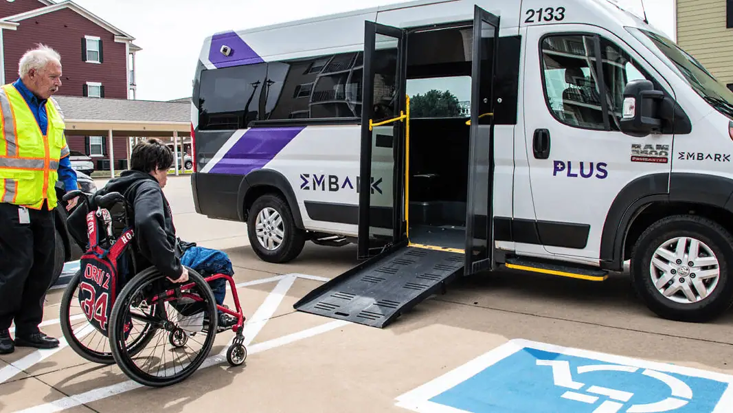 EMBARK paratransit 2 Caregiver in a safety vest helps a person in a red wheelchair toward a white shuttle van with a lowered ramp in a parking lot.