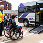 Caregiver in a safety vest helps a person in a red wheelchair toward a white shuttle van with a lowered ramp in a parking lot.