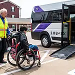 EMBARK driving accessibility, assistance for public transit in OKC Caregiver in a safety vest helps a person in a red wheelchair toward a white shuttle van with a lowered ramp in a parking lot.