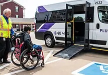 EMBARK driving accessibility, assistance for public transit in OKC Caregiver in a safety vest helps a person in a red wheelchair toward a white shuttle van with a lowered ramp in a parking lot.
