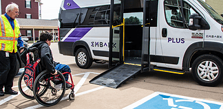 Caregiver in a safety vest helps a person in a red wheelchair toward a white shuttle van with a lowered ramp in a parking lot.