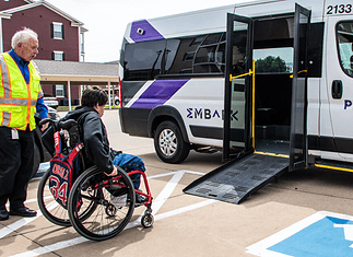 EMBARK driving accessibility, assistance for public transit in OKC Caregiver in a safety vest helps a person in a red wheelchair toward a white shuttle van with a lowered ramp in a parking lot.