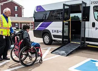 EMBARK driving accessibility, assistance for public transit in OKC Caregiver in a safety vest helps a person in a red wheelchair toward a white shuttle van with a lowered ramp in a parking lot.