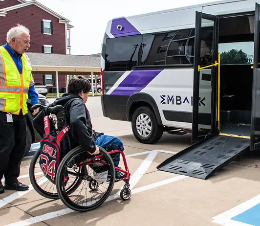 EMBARK driving accessibility, assistance for public transit in OKC Caregiver in a safety vest helps a person in a red wheelchair toward a white shuttle van with a lowered ramp in a parking lot.