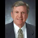 PRESS RELEASE: Former Oklahoma City Mayor Guy Liebmann passes away Professional head-and-shoulders portrait of a smiling middle-aged man in a dark suit and patterned tie against a muted blue-gray background.