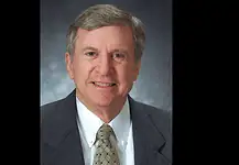 PRESS RELEASE: Former Oklahoma City Mayor Guy Liebmann passes away Professional head-and-shoulders portrait of a smiling middle-aged man in a dark suit and patterned tie against a muted blue-gray background.