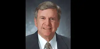 Professional head-and-shoulders portrait of a smiling middle-aged man in a dark suit and patterned tie against a muted blue-gray background.