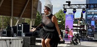 Drag performer wearing a tall silver crown and black sequined outfit performing on an outdoor stage at the Two Spirit Festival