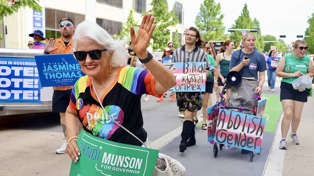 Older woman with white hair in a rainbow shirt and sunglasses waves at a Pride parade, holding a green campaign sign for Cyndi Munson for Governor.