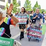 Older woman with white hair in a rainbow shirt and sunglasses waves at a Pride parade, holding a green campaign sign for Cyndi Munson for Governor.