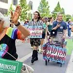 Oklahoma Pride Alliance looks beyond Pride Month Older woman with white hair in a rainbow shirt and sunglasses waves at a Pride parade, holding a green campaign sign for Cyndi Munson for Governor.