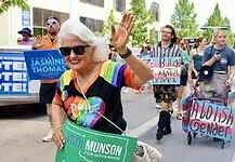 Oklahoma Pride Alliance looks beyond Pride Month Older woman with white hair in a rainbow shirt and sunglasses waves at a Pride parade, holding a green campaign sign for Cyndi Munson for Governor.